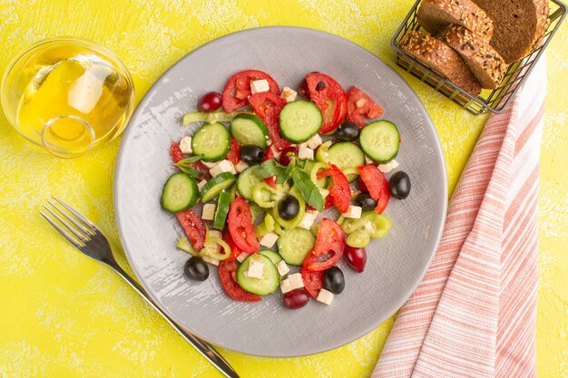 Вид сверху салат из свежих овощей с нарезанными огурцами помидоры оливковое внутри тарелка с маслом и хлебом на желтой поверхности овощная еда салат цвет еды