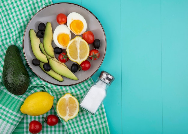 Top view of fresh vegetables on bowl with avocados lemon olives tomatoes with salt shaker on green checked tablecloth on blue with copy space