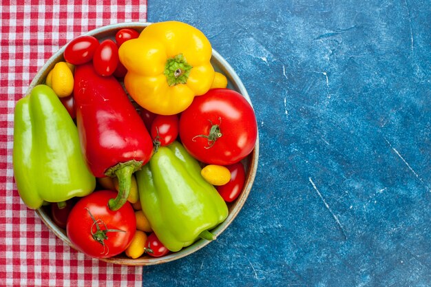 Top view fresh vegetables cherry tomatoes different colors bell peppers tomatoes in bowl on red and white checkered tablecloth on blue table with copy space