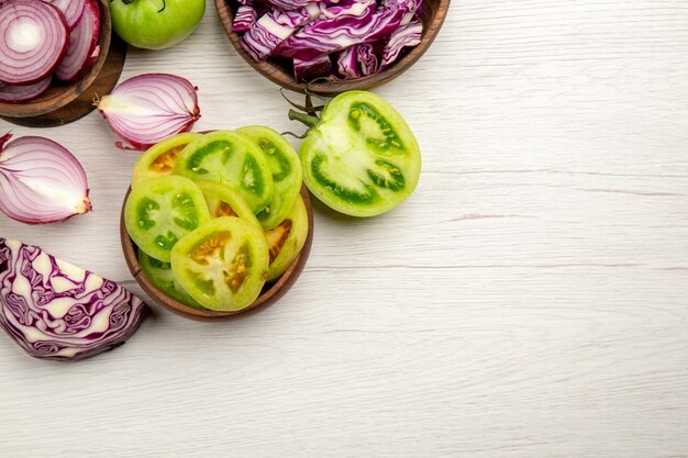 Top view fresh vegetables cut green tomatoes cut red cabbage cut onion in bowls on white wooden surface with free place