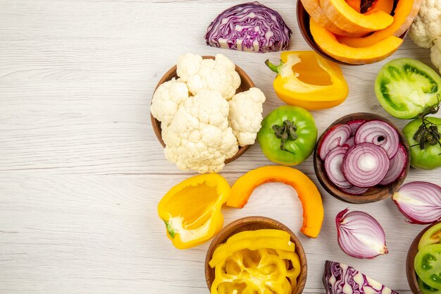 Top view fresh vegetables cut green tomatoes cut red cabbage cut onion cut pumpkin cauliflower cut bell pepper in bowls on white wooden surface free space