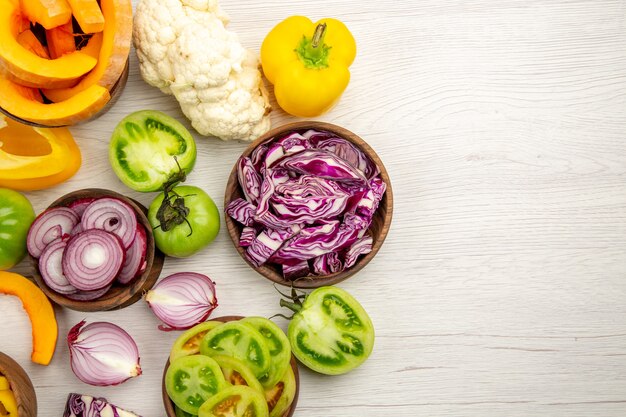 Top view fresh vegetables cut green tomatoes cut red cabbage cut onion cut pumpkin cauliflower cut bell pepper in bowls on white wooden surface with copy space