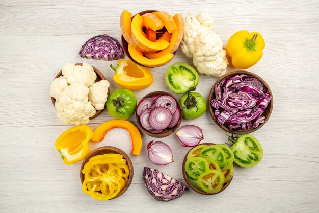 Top view fresh vegetables cut green tomatoes cut red cabbage cut onion cut pumpkin cauliflower cut bell pepper in bowls on white wooden table