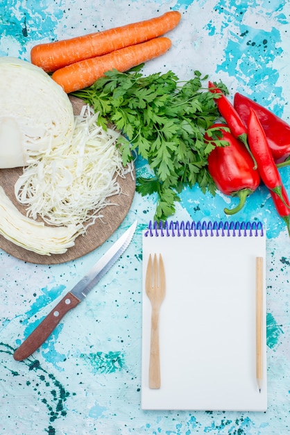 Top view of fresh vegetables greens sliced cabbage carrots and peppers with notepad on bright-blue desk, food meal vegetable lunch healthy