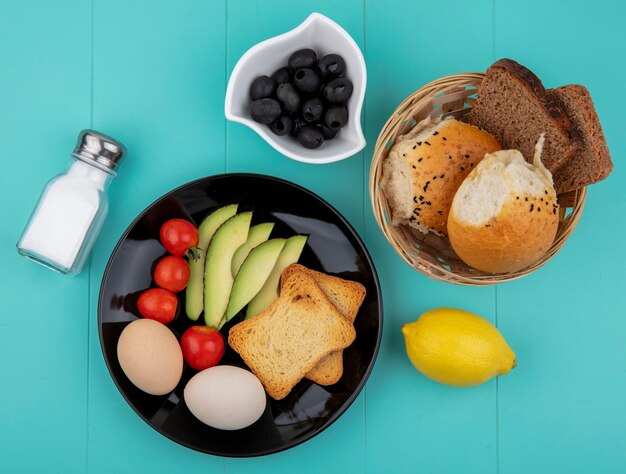 Top view of fresh vegetables like avocado slices tomatoes olives eggs on black plate with a bucket of breads lemon on blue