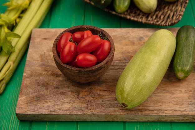 Вид сверху на свежие овощи, такие как кабачки огурцов на деревянной кухонной доске со сливовыми помидорами на деревянной миске с сельдереем, изолированные на зеленой деревянной стене