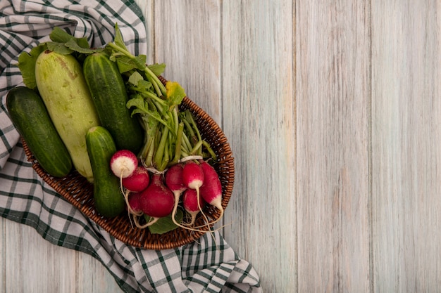 Top view of fresh vegetables such as cucumbers zucchinis and radishes on a bucket on a checked cloth on a grey wooden wall with copy space