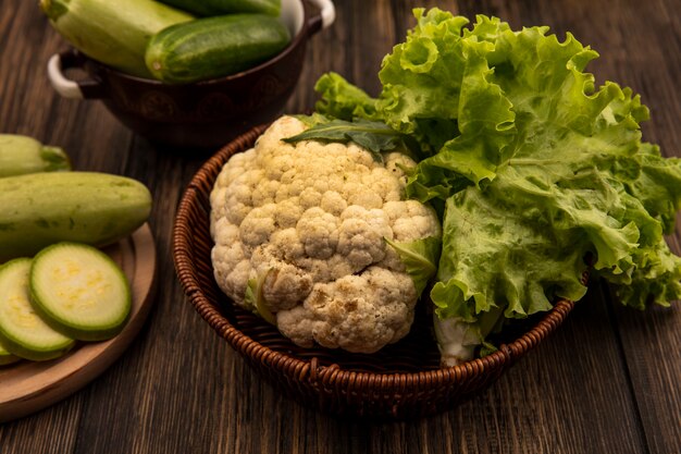 Top view of fresh vegetables such as lettuce and cauliflower on a bucket with chopped zucchinis on a wooden kitchen board with cucumbers on a bowl on a wooden surface