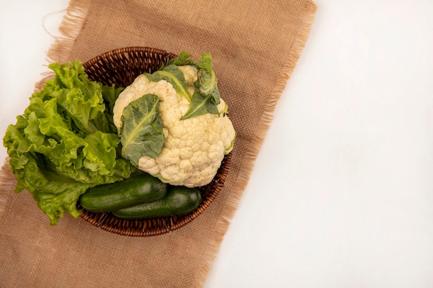 Top view of fresh vegetables such as lettuce cauliflower and cucumbers on a bucket on a sack cloth on a white background with copy space