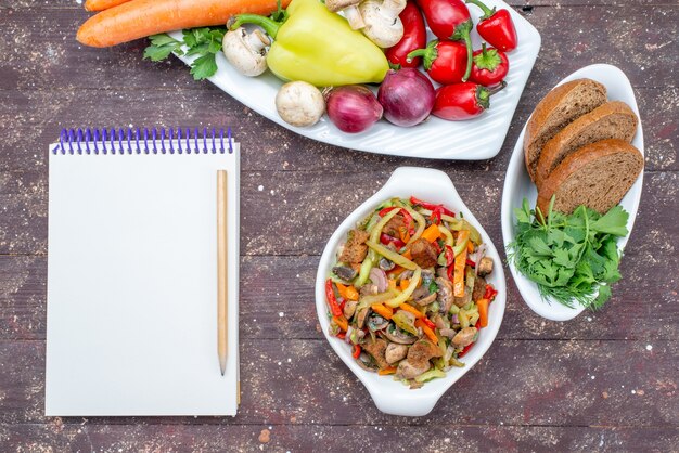 Top view of fresh vegetables with mushrooms inside plate with bread loafs and greens notepad on brown, vegetable food meal mushroom