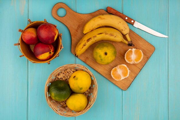 Top view of fresh whole and half tangerines on a wooden kitchen board with bananas with knife with peaches on a bucket on a blue wooden wall