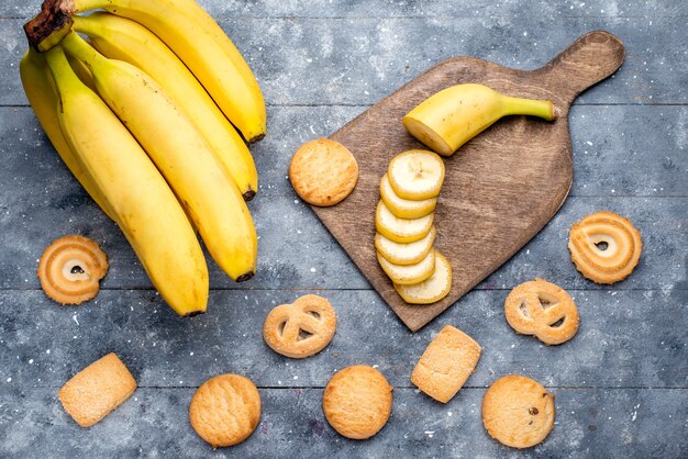 top view of fresh yellow bananas sliced and whole along with cookies on grey, fresh fruit berry