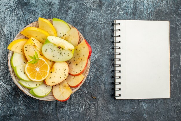 Top view of fresh yellow green red apple slices on a white plate and spiral notebook on gray background