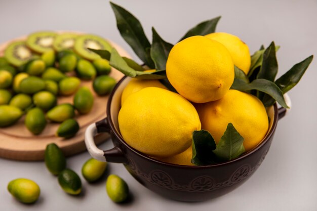 Вид сверху свежих желтых лимонов на миске с кинканами и ломтиками киви на деревянной кухонной доске на белой стене