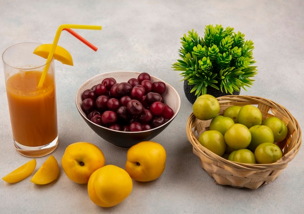 Top view of fresh yellow peaches with fresh peach juice with red cherries on a bowl with green cherry plums on a bucket on a white background