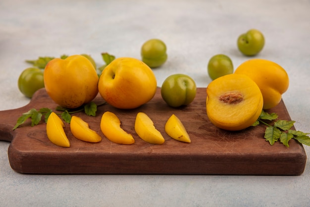 Вид сверху свежих желтых персиков с дольками на деревянной кухонной доске с зелеными алычами, изолированными на белом фоне