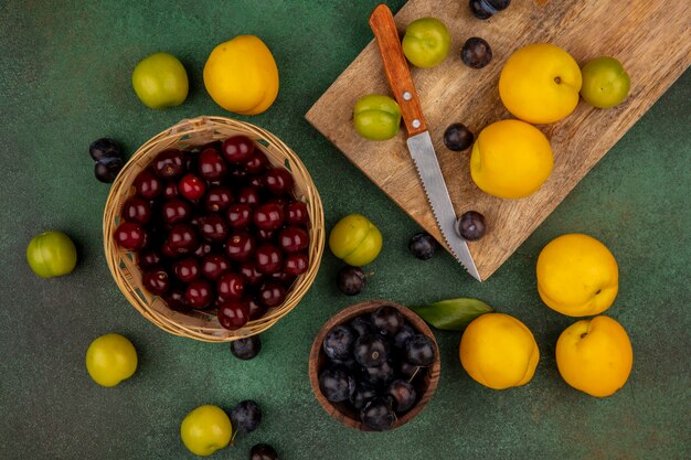 Top view of fresh yellow peaches on a wooden kitchen board with green cherry plums with red cherries on a bucket on a green background