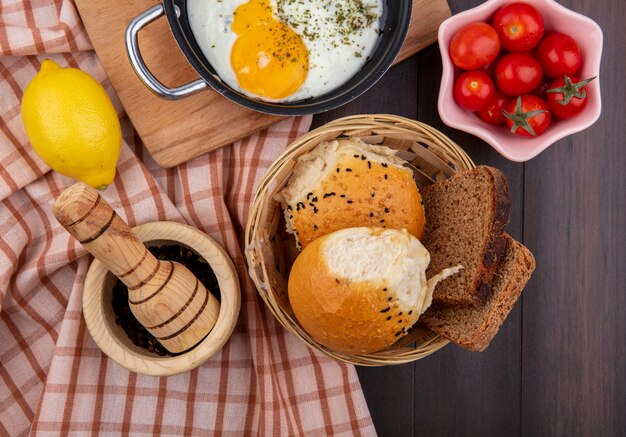 Top view of fried egg in a frying pan with a bucket of breads tomatoes lemon on checked tablecloth