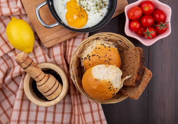 Вид сверху жареного яйца на сковороде с ведром хлеба, помидоров, лимона на проверенной скатерти