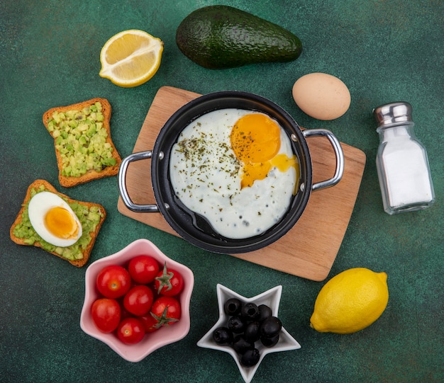 Free photo top view of fried egg in a frying pan on wood kitchen board with tomtoes lemon black olives on green