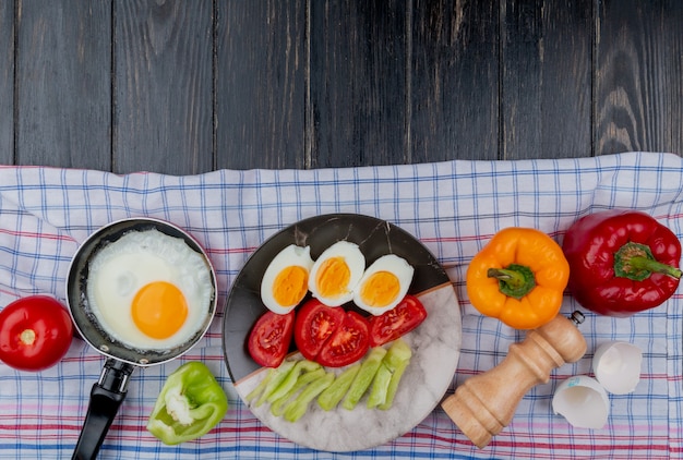 Free photo top view of fried egg with boiled halved eggs on a plate with tomato slices and bell pepper slices on a checked tablecloth on a wooden background with copy space