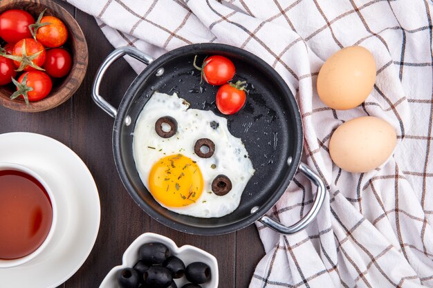 Top view of fried egg with tomatoes and olives in pan and eggs on plaid cloth with bowls of tomato and olive cup of tea on wood