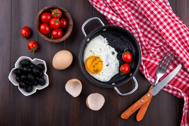 Top view of fried egg with tomatoes in pan and fork with knife on plaid cloth and egg with shell and bowls of tomato and olive on wood