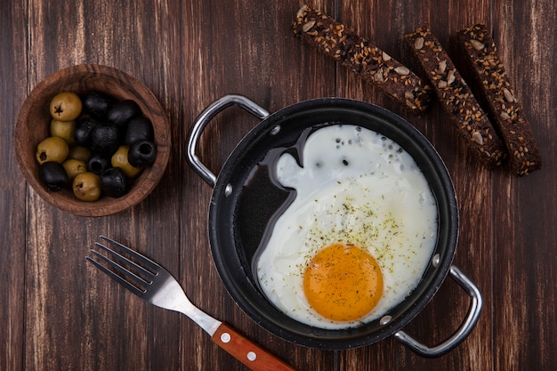 Top view fried eggs in a frying pan with    slices of black bread and olives with fork on wooden background