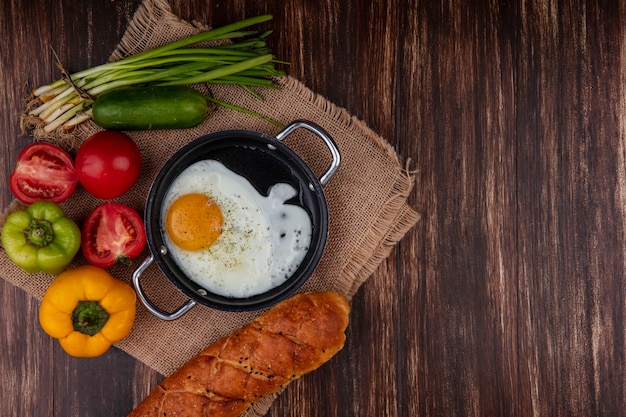 Top view fried eggs in a pan with green onions  tomatoes  cucumber  bell peppers and a loaf of bread  on a beige napkin  on a wooden background