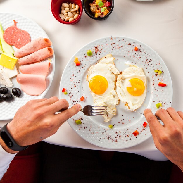 白いテーブルクロス、オリーブ、チーズ、ハム、ナッツ、フォークとナイフの朝食を持つ男の砂糖漬けの果物の手でテーブルの上のテーブルに目玉焼き