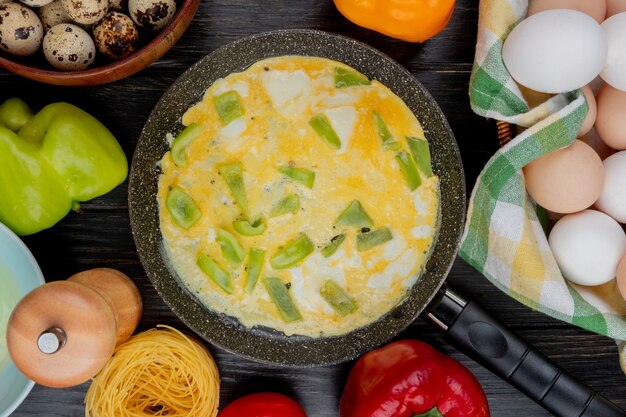 Top view of fried eggs with chopped slices of green bell pepper on a frying pan with fresh chicken eggs on a wooden background