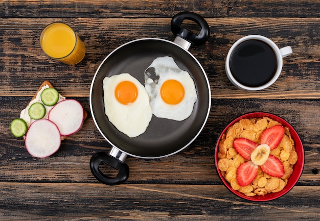 Top view of fried eggs with toasts, cornflakes and drinks on dark wooden surface horizontal