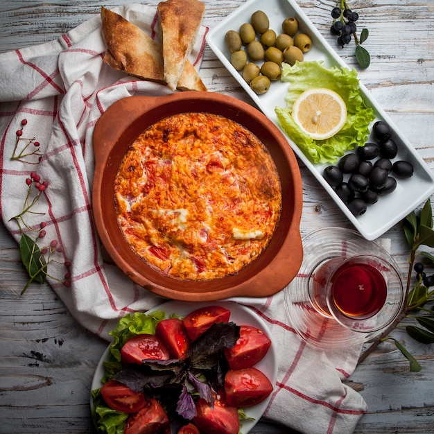 Top view fried eggs with tomatoes with glass of tea and assorted olives and tomatoes in clay plate