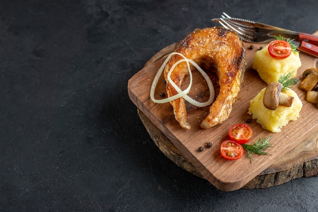 Top view of fried fish meal with mushrooms vegetables cheese and cutlery set on wooden board on the left side on black distressed surface