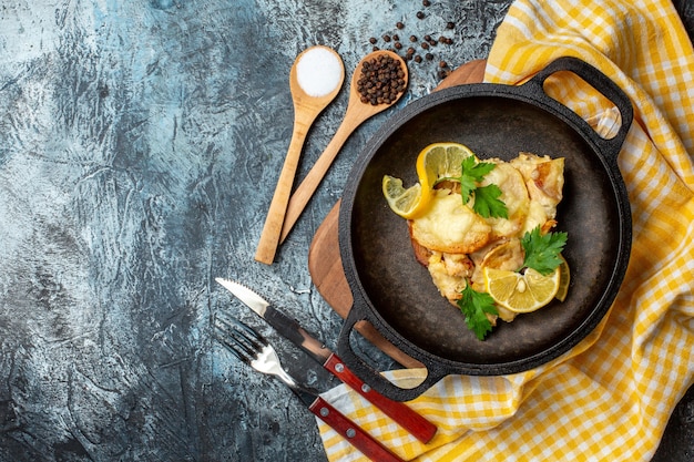 Top view fried fish in pan with lemon and parsley salt and pepper in wooden spoons