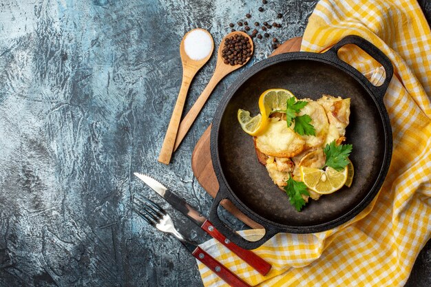 Top view fried fish in pan with lemon and parsley salt and pepper in wooden spoons