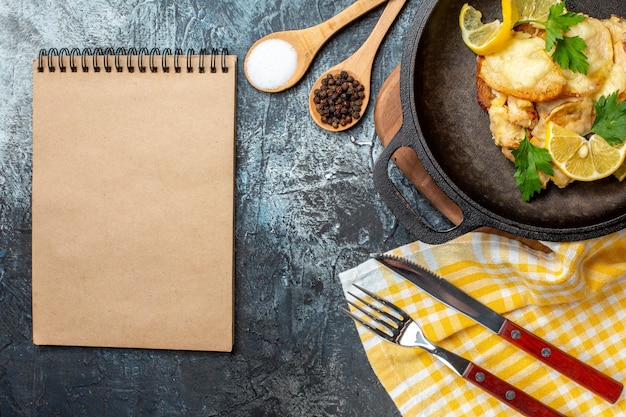 Foto gratuita vista dall'alto pesce fritto in padella con limone e prezzemolo spezie in una ciotola e cucchiai di legno forchetta e coltello blocco note su sfondo grigio