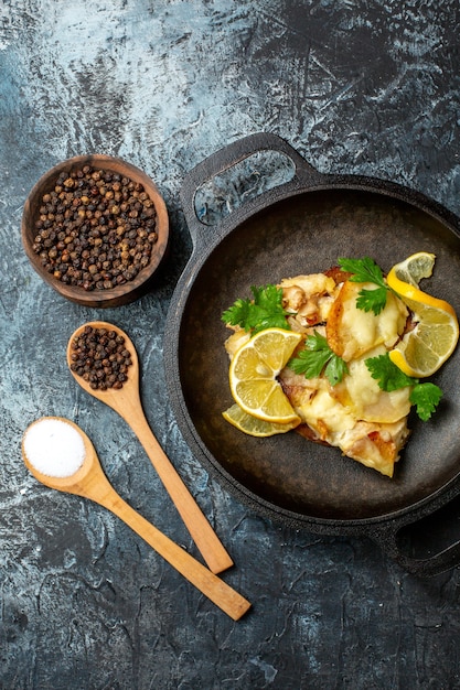 Top view fried fish in pan with lemon and parsley spices in wooden spoons black pepper in bowl on grey background