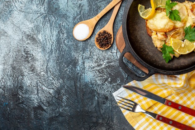 Вид сверху жареная рыба на сковороде со специями из лимона и петрушки в деревянных ложках, вилке и ноже на сером фоне