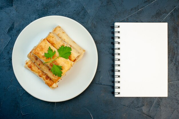 Top view of fried lavash wraps on a plate served with green and spiral notebook on dark background