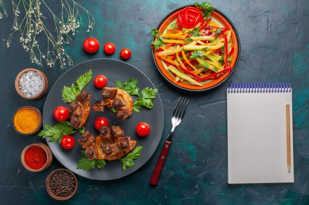 Top view fried meat slices with sliced vegetable salad notepad and seasonings on dark-blue desk vegetable food meat health meal