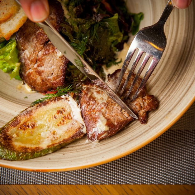 Top view fried meat with fried zucchini and fork and knife and greens in round plate