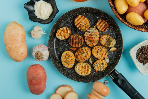 Top view of fried potato slices in frying pan with uncooked ones in basket mayonnaise garlic salt black pepper on blue