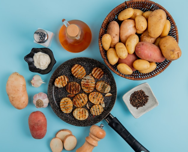 Top view of fried potato slices in frying pan with uncooked ones in basket mayonnaise garlic salt black pepper and butter on blue