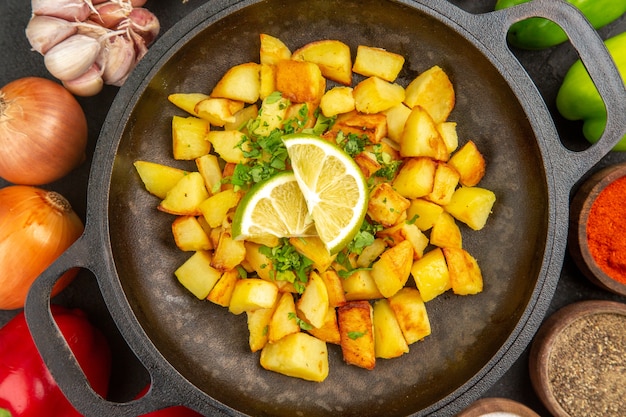 Вид сверху жареный картофель внутри сковороды с разными приправами и овощами на темном фоне