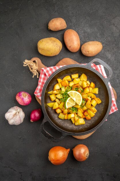 Вид сверху жареный картофель внутри сковороды со свежими овощами на темном фоне