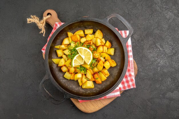Top view fried potatoes inside pan with greens and lemon on a dark background