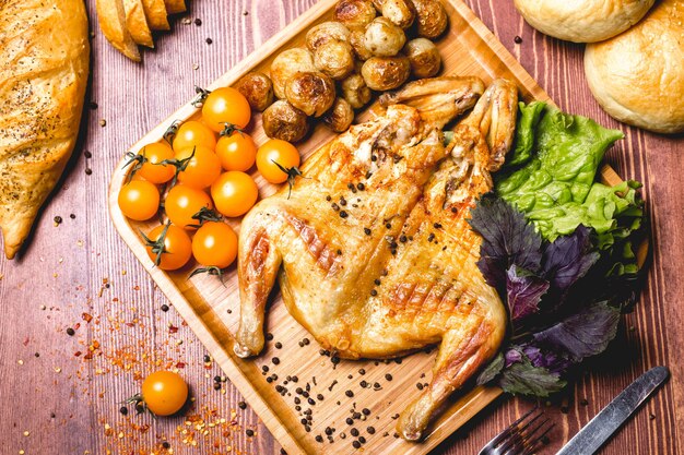 Top view fried tobacco chicken with baked potatoes and yellow ceri tomatoes with salad leaves on a board