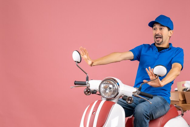 Top view of frigthened courier guy wearing hat sitting on scooter delivering orders on pastel peach background