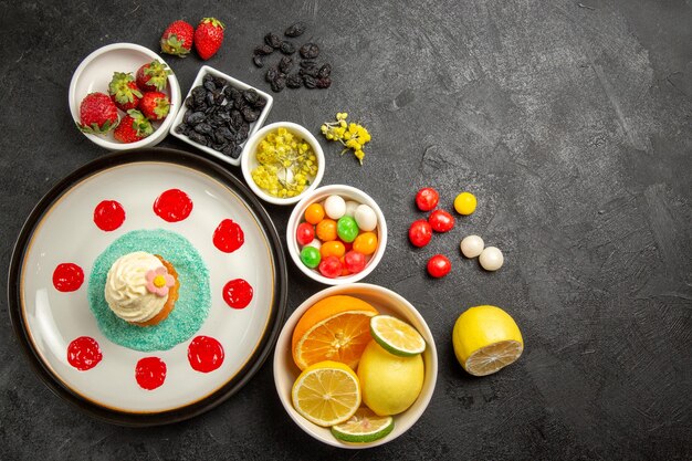 Top view from afar berries and fruits cake with sauces and cream next to the white bowls of strawberries limes lemons oranges and colorful sweets on the table